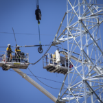 Men atop a cherry picker attaching wires to a pylon with the use of a crane
