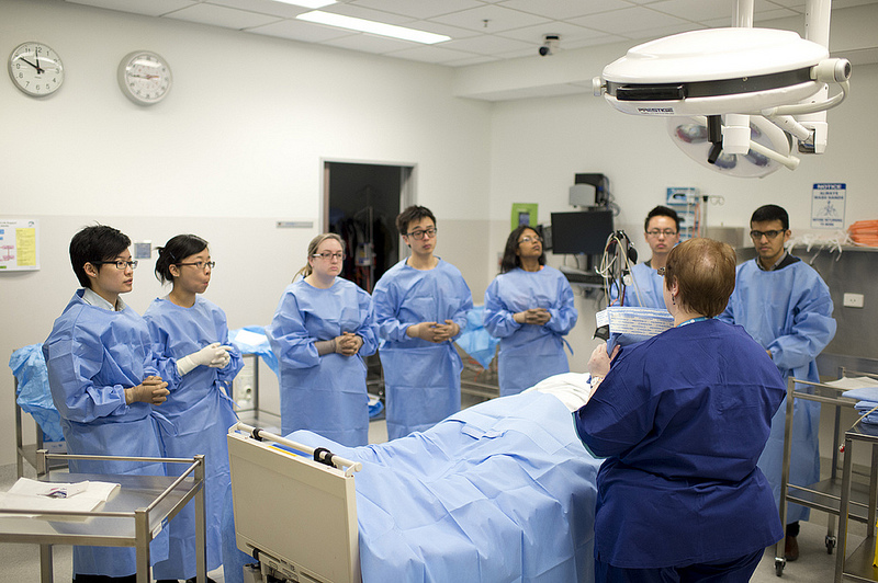 Students gathered around a patient in a hospital bed whilst instructed by a doctor.